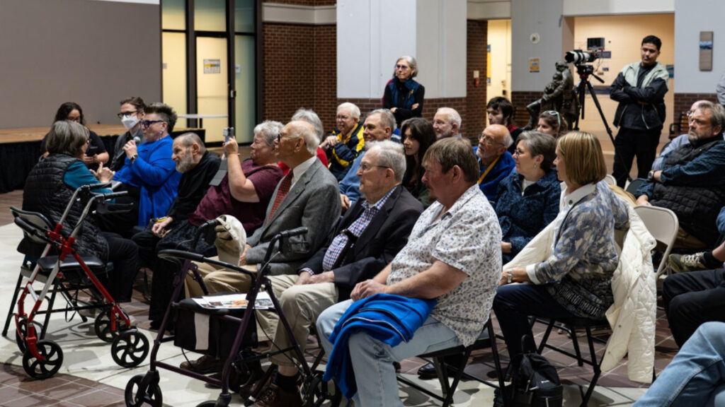 A photo of a seated crowd of at least twenty people in a large room. Many appear to be senior. Two walkers are in the front row. In the back is a standing person and a cameraperson with camera.