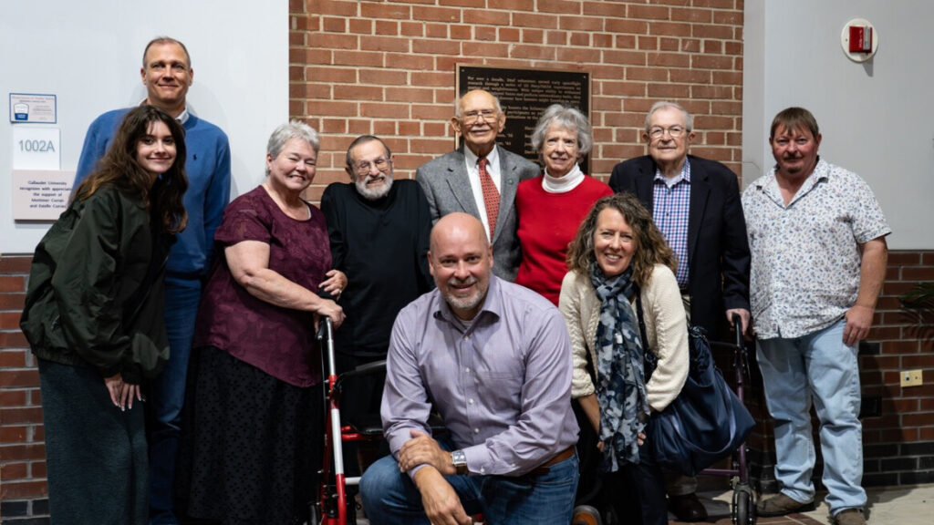A photo of ten people posing and smiling for the camera. They stand and kneel in front of a brick wall with a plaque. Several appear to be older.
