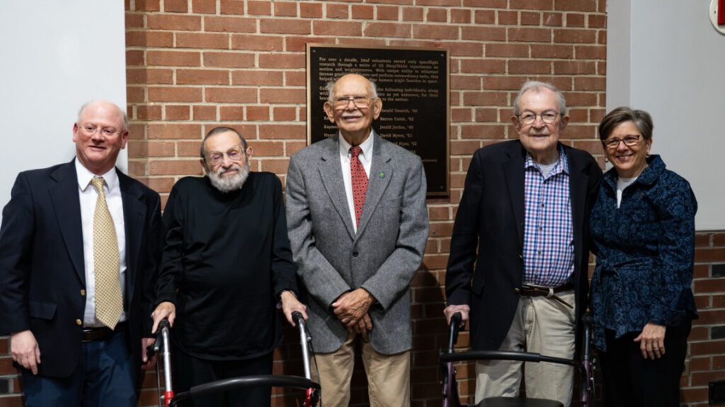 A photo of five people smiling for the camera, in front a plaque on a brick wall. The three in the middle are senior. Two (second and fourth from left) have walkers. Everybody is formally dressed.