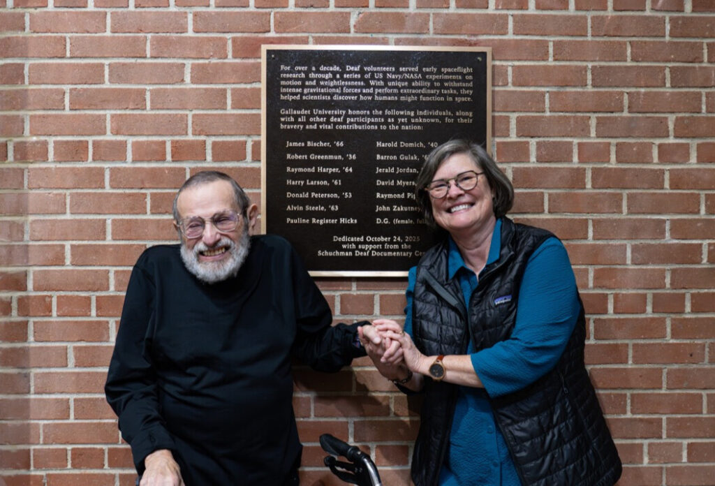 A photo of two people smiling for the camera, in front a plaque on a brick wall. The person on the right is warmly clasping the hand of the person on the left, who has a walker.