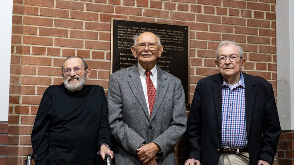 A photo of three seniors smiling for the camera, in front a plaque on a brick wall. Two (first and third from left) have walkers. Everybody is formally dressed.