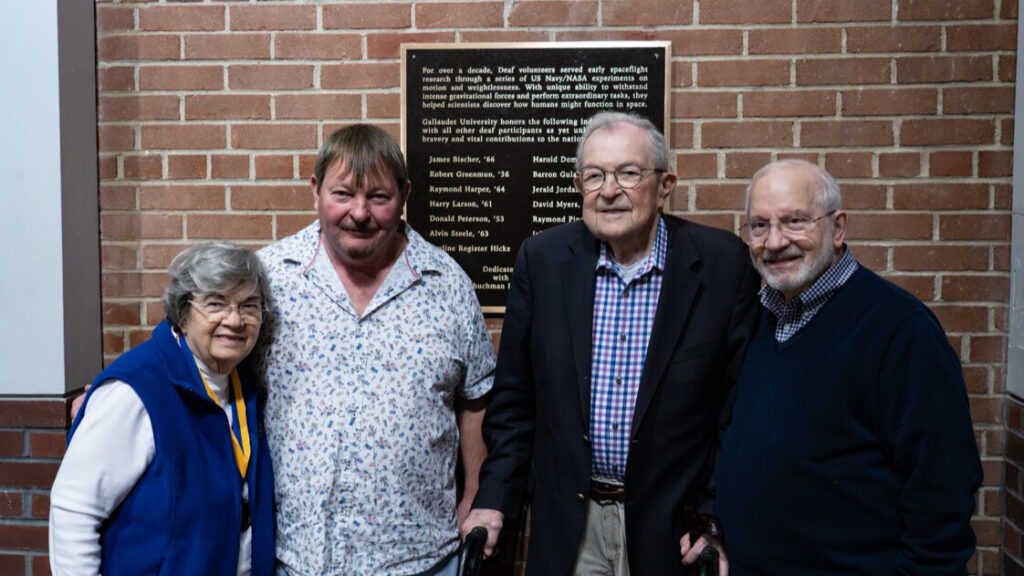 A photo of four people smiling for the camera, in front a plaque on a brick wall. The person third from left has a walker.