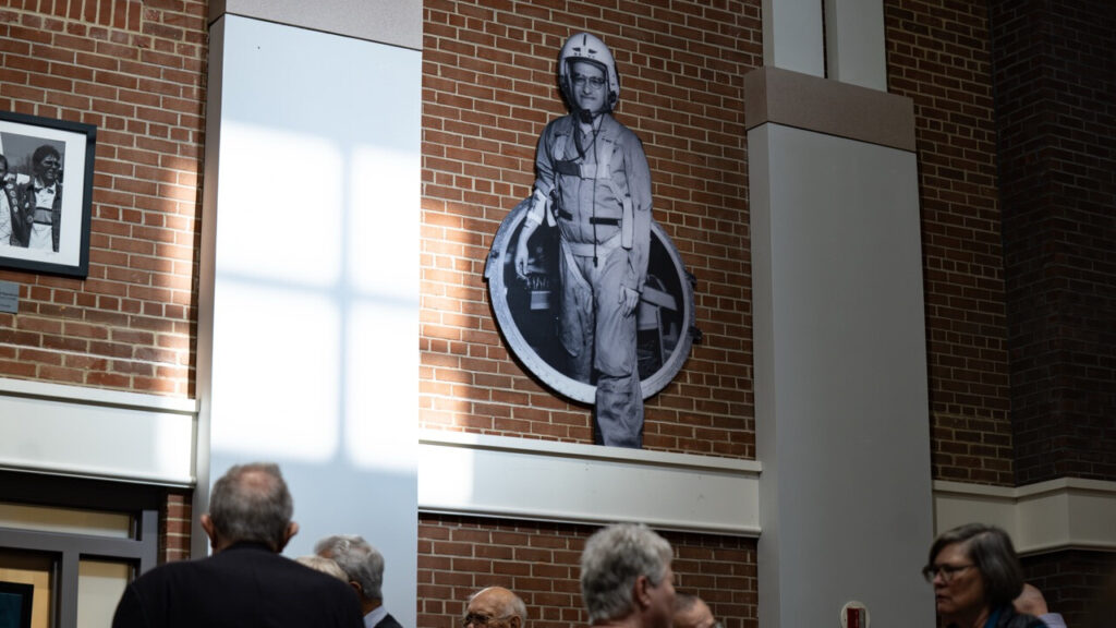 A photo looking up a wall in a large room. In the center is a brick panel with a black and white cutout sign. The sign shows what appears to be an astronaut emerging from a hole-shaped vehicle door. At the bottom of the image, out-of-focus heads are visible in the room.