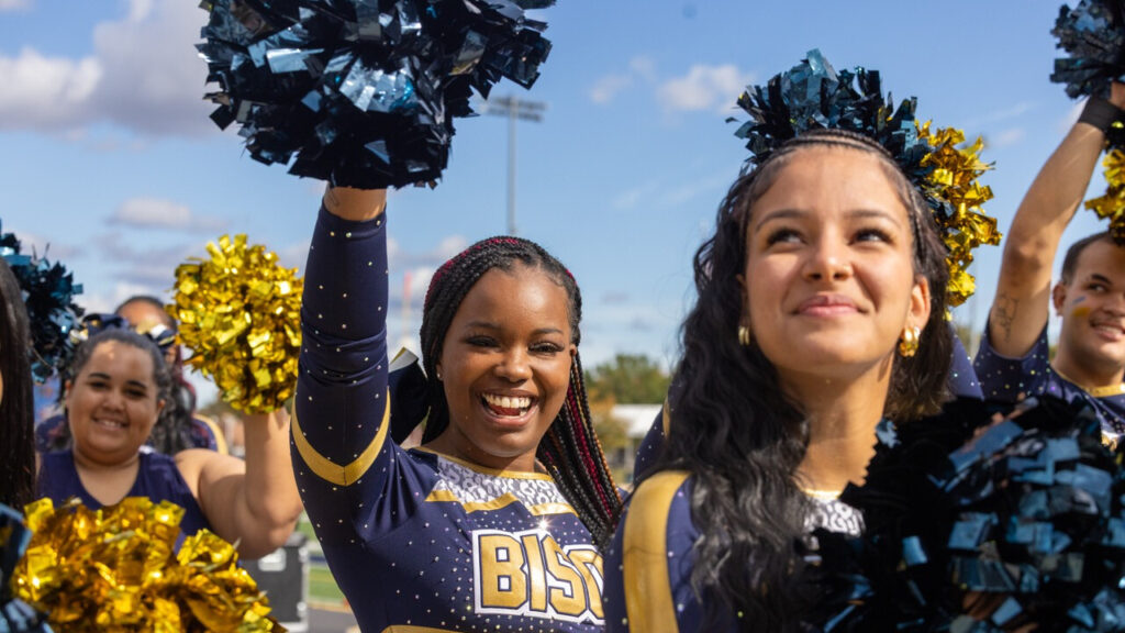 A photo of three smiling cheerleaders in blue and gold uniforms. In the center of the image, a cheerleader raises a blue pompom with their right arm. They have stylish braids over their left shoulder. The obscured name on their uniform can be understood to say "Bison".