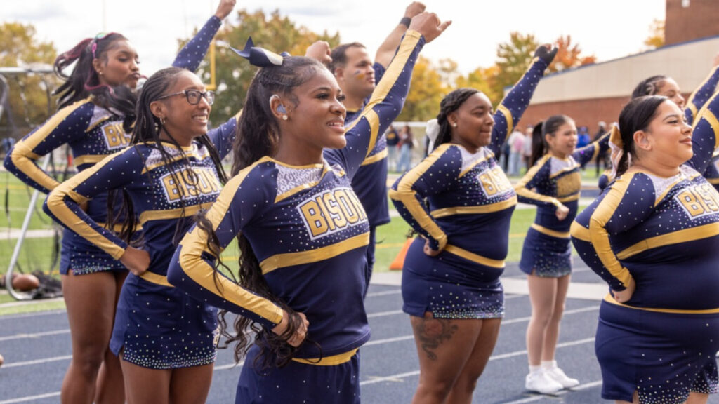 A photo at an angle of eight cheerleaders striking a pose for an unseen crowd to the right. They are wearing matching blue and gold uniforms with skirts. They are standing on a track, with a field in the background.