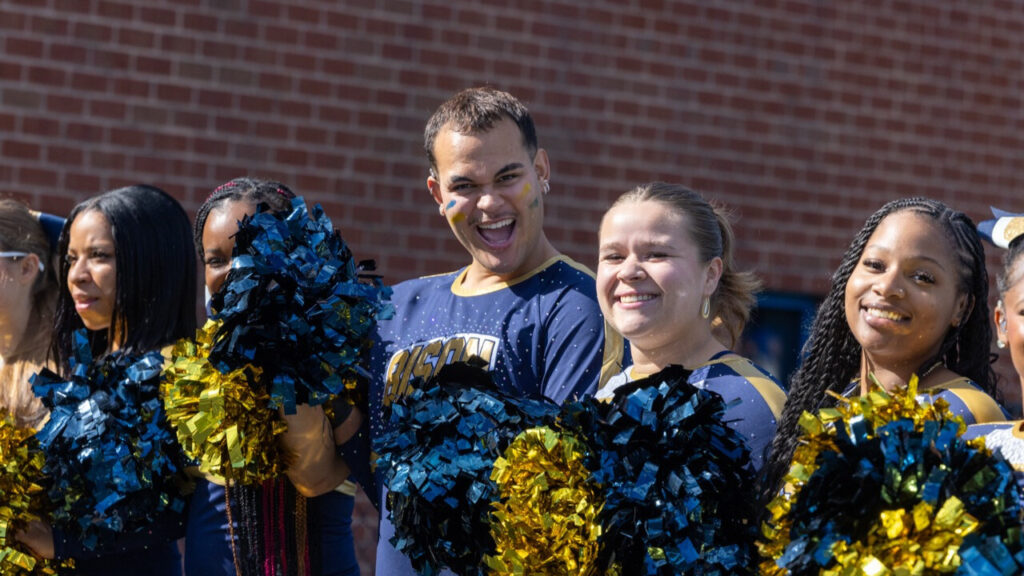 A photo of five smiling cheerleaders in blue and gold uniforms. Prominent in the center is a taller, masculine-presenting cheerleader looking directly at the camera with cheer.