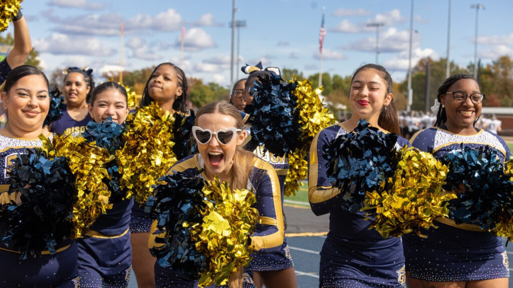Seven cheerleaders in blue and gold smile and shake pompoms for the camera. Fifth from the left, in the center of the image, a cheerleader catches the eye with white sunglasses in the shape of hearts. They are cheering intensely.