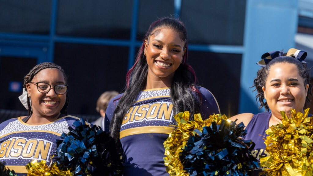 A photo of three cheerleaders in blue and gold smiling for the camera. The cheerleader in the middle is much taller than the other two. Their uniforms say "Bison" and they hold blue and gold pompoms.