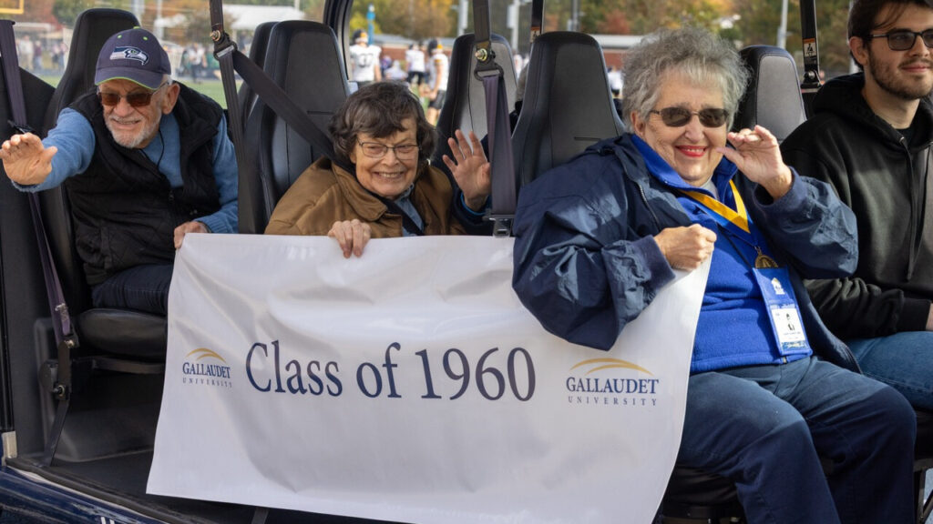 A photo of three seniors riding in a golf cart, smiling at the camera. They are holding a white banner that says "Class of 1960" with the Gallaudet University logo.