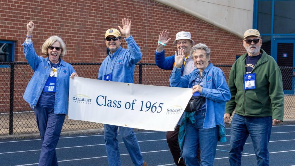 A photo of five seniors walking on an athletic track, smiling and waving for the camera. The two in front are holding a white banner that says "Class of 1965" with the Gallaudet University logo.