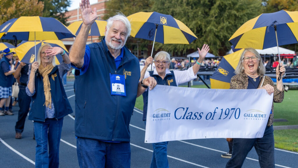 A photo of four seniors walking on an athletic track, smiling and waving for the camera. Three are holding blue and yellow umbrellas. The two in front are holding a white banner that says "Class of 1970" with the Gallaudet University logo.