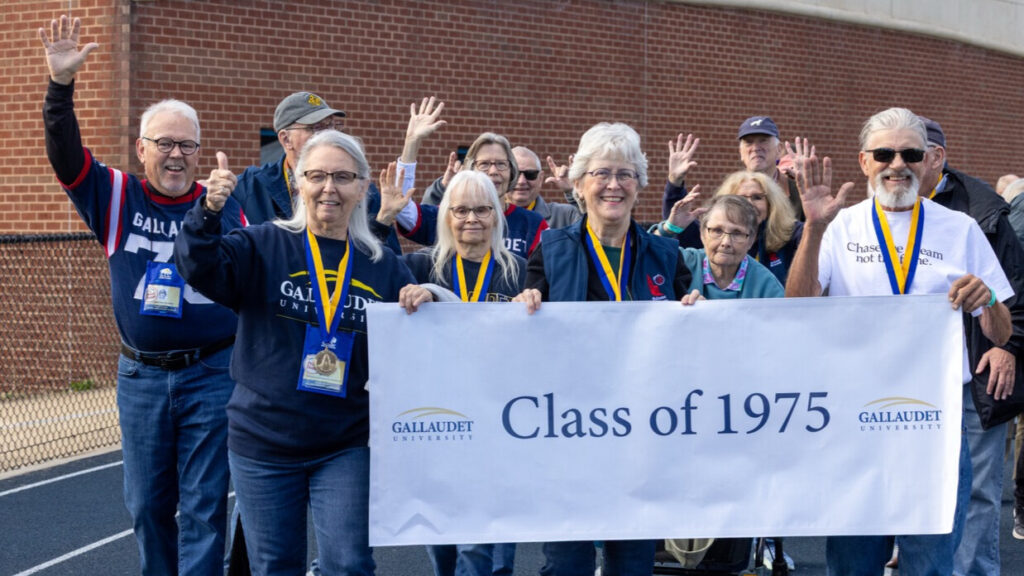 A photo of at least twelve seniors walking on an athletic track, smiling and waving for the camera. Many are wearing dark blue. Three in the front are holding a white banner that says "Class of 1975" with the Gallaudet University logo.
