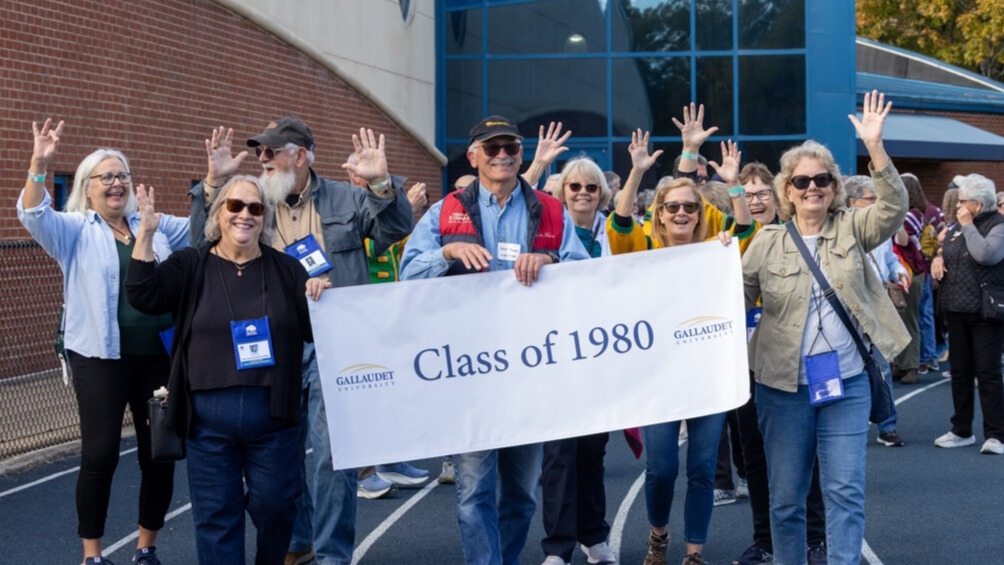 A photo of at least eight seniors walking on an athletic track, smiling and waving for the camera. Three in the front are holding a white banner that says "Class of 1980" with the Gallaudet University logo.