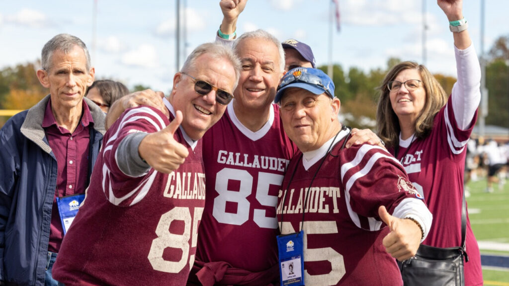 A photo of three seniors smiling for the camera. They wear matching green and white football jerseys that say "Gallaudet" and have the number 85. The person in the middle has their hands around the other two, who give a thumbs up. Two other people are visible in the background.