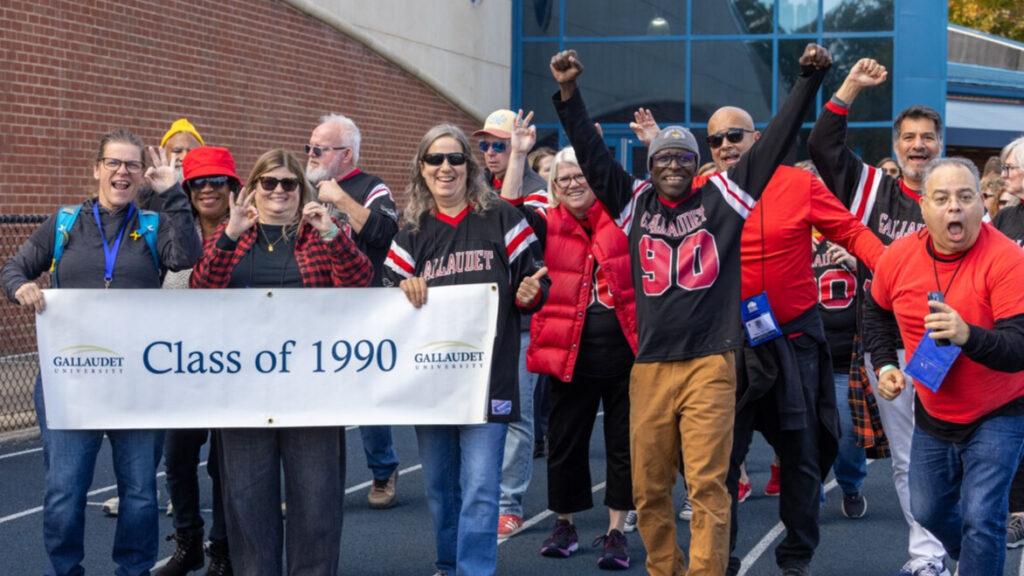 A photo of at least eleven people walking on an athletic track, smiling and cheering for the camera. Many are wearing black and red. On the left, three are holding a white banner that says "Class of 1990" with the Gallaudet University logo. On the right, two others make enthusiastic gestures.