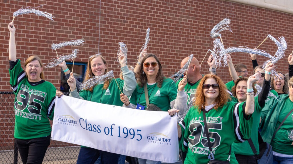 A photo of at least ten people walking on an athletic track, smiling and cheering for the camera. Many are wearing green apparel and holding silver streamers Two in front hold a white banner that says "Class of 1995" with the Gallaudet University logo. On the left, a particularly enthusiastic individual has their right arm fully extended, holding a streamer.