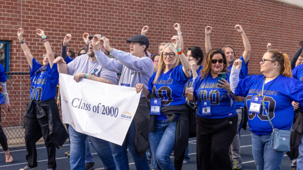 A photo of at least ten people walking on an athletic track, smiling and cheering for an unseen crowd to the left. Many are wearing black and sky blue football jerseys and raising their hands with a double zero. Three in front are holding a white banner that says "Class of 2000" with the Gallaudet University logo.