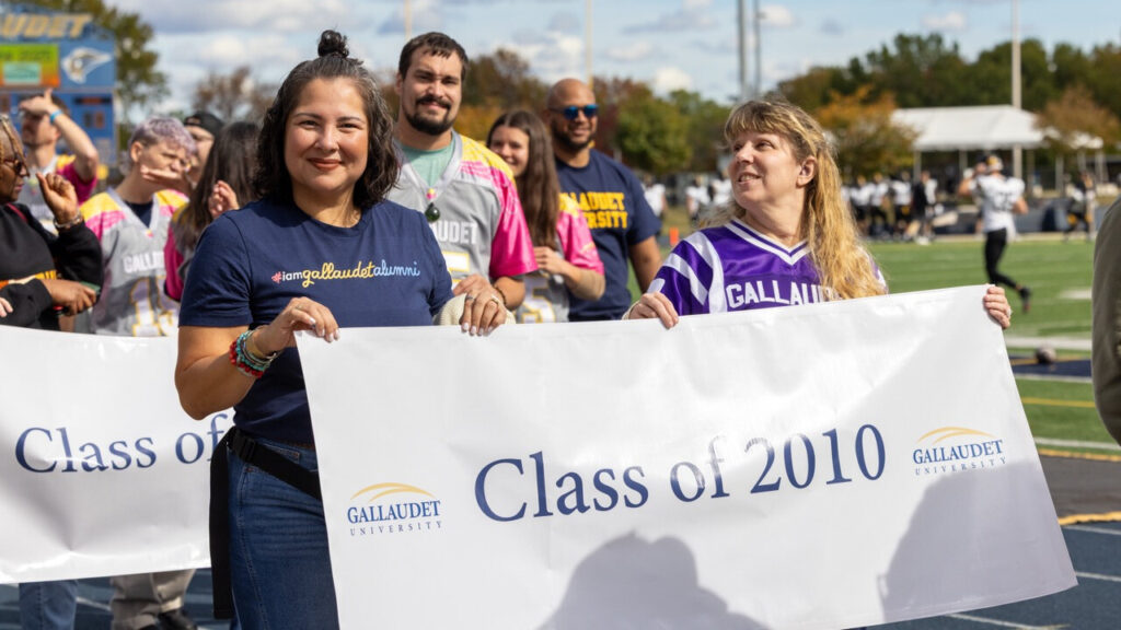 A photo of at least either people walking on an athletic track. In the foreground, two hold a white banner that says "Class of 2010" with the Gallaudet University logo. One smiles for the camera and the other looks offscreen to the left.