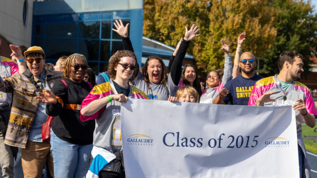 A photo of at least nine people walking on an athletic track, smiling and cheering for the camera. In front, two in gray baseball jerseys hold a white banner that says "Class of 2015" with the Gallaudet University logo. In the back are four extended arms in celebration.