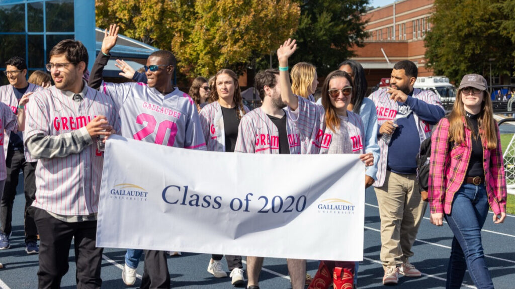 A photo of at least eleven people walking on an athletic track, smiling and cheering for the camera. In front, two in gray baseball jerseys hold a white banner that says "Class of 2020" with the Gallaudet University logo. In the back, more gray jerseys are visible.