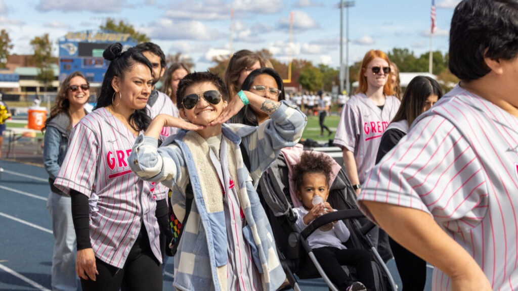 A photo of at least ten people walking on an athletic track. Some are wearing gray baseball jerseys. Prominent in the left center is a person in sunglasses smiling broadly with their hands under their chin. To their left in the background is a small child in a stroller.