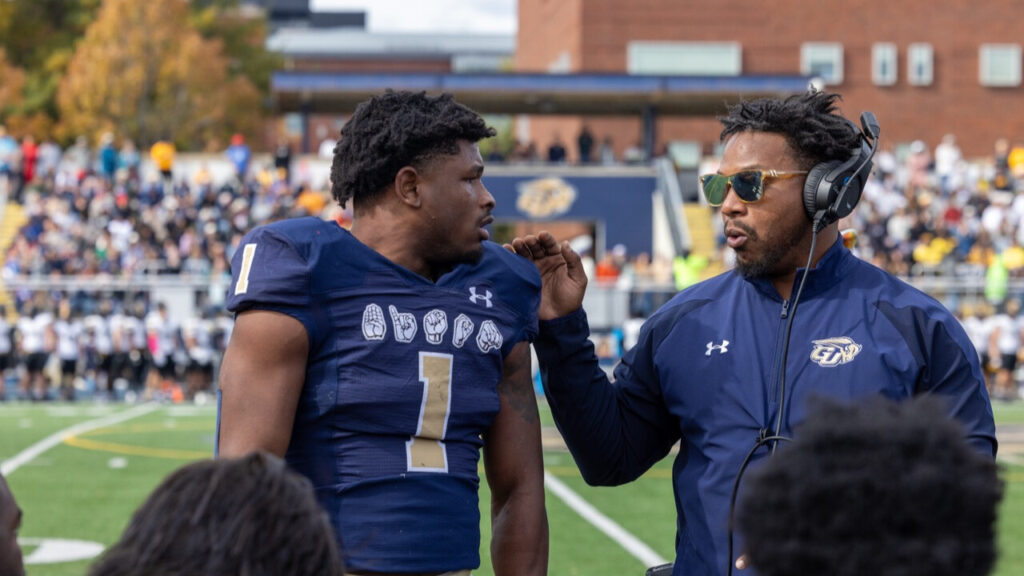 A photo of a football player (left) and coach (right) looking at each other on the sideline of a football game. They are wearing dark blue. The player's uniform has "Bison" in ASL handshapes. The coach has sunglasses and a headset.
