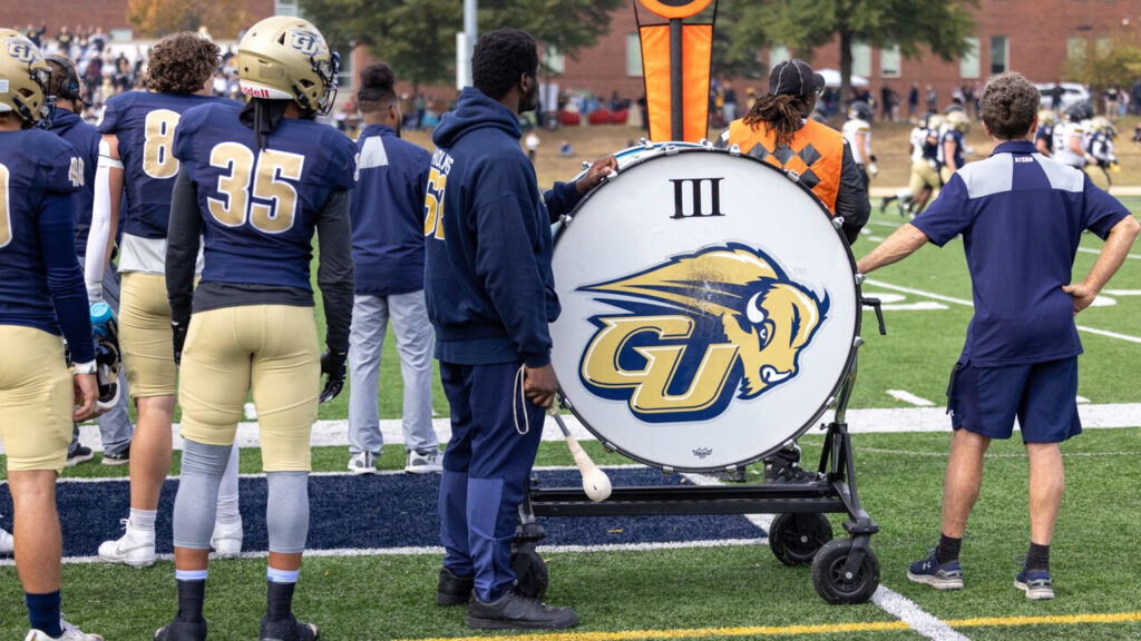 A photo behind the sideline of a football game. Prominent on the right side is a large drum with the Gallaudet Athletics logo. To the left of the drum is a person in blue garb holding a large mallet. On the left of the image are football players in blue and gold looking at the field.