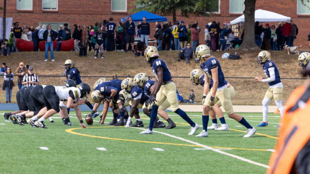 A photo of a football game from the sidellines. On the right, a team in blue and gold gets ready to hike the ball. On the left, a team in black and white prepares to defend. In the background are observers on a small hill.