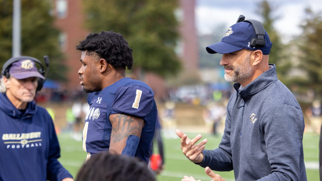 A photo of two football coaches (background left and foreground right) and a player (foreground center). The player is facing left. Their uniform is blue and gold. The coach in the foreground is also facing right, with a blue hat and headset. They have an intense look.