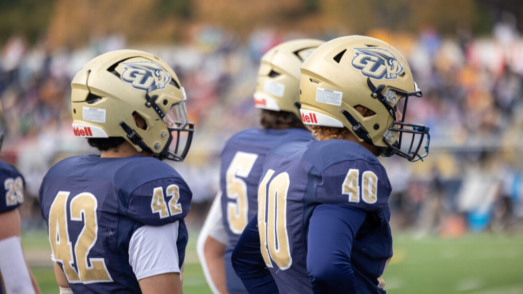 A photo at an angle of three football players in watching a football game offscreen to the right. They have gold helmets with the Gallaudet Athletics logo, and blue jerseys with gold numbers.