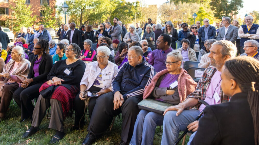 A photo of a crowd of over fifty people on grass on a sunny day, looking to the left at an unseen presentation. The people in the front are seated, while people in the back stand. Trees are visible in the far background.