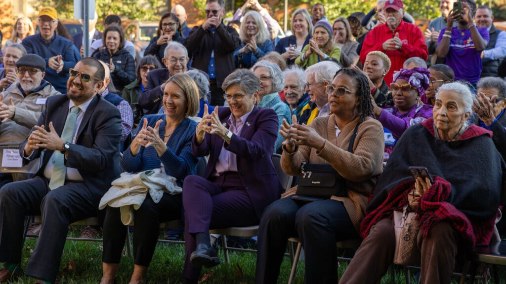 A photo of a crowd of over fifty people on grass in the shade on a sunny day, looking to the left at an unseen presentation and copying an ASL sign for "necessity". The people in the front are seated, while people in the back stand.