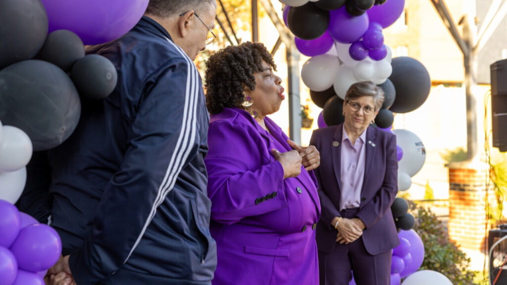 A photo of a presentation outdoors. Prominent in the center, somebody presents in a purple pantsuit. To their right in the background, a person watches with their hands clasped at their waist. In the foreground, another person watches with their hands clasped behind their back. Around them is an arc of purple, white, and black balloons.