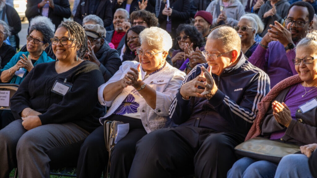 A photo of a crowd of seated people in the shade on a sunny day, looking to the left at an unseen presentation and copying an ASL sign for "necessity".