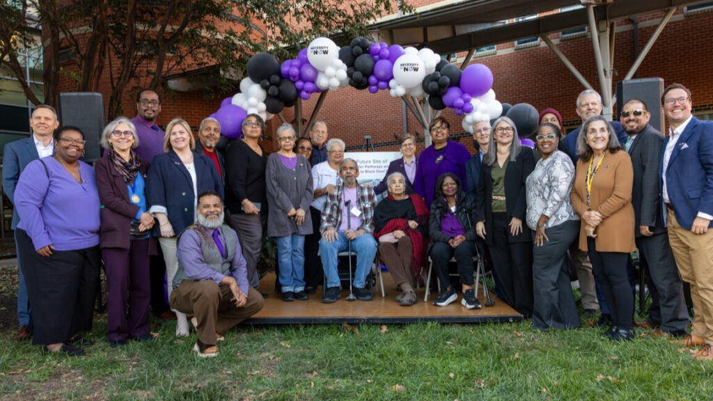 A photo of 25 people posing around a small outdoor stage, smiling for the camera. The stage has a decorative arc of purple, white, and blue balloons. Three people on the stage are seated. The rest stand around and on the stage, other than one kneeling individual.