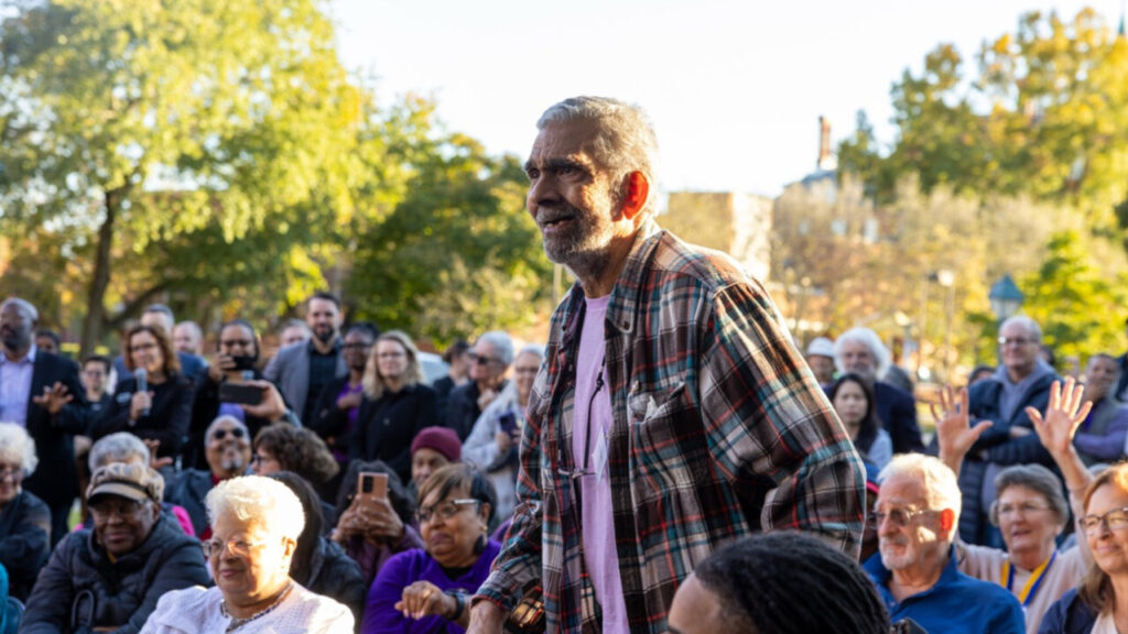 A photo of an older person standing up amongst seated people, outdoors in the shade on a sunny day. They are looking an unseen presentation to the left. More people in the background are standing. Trees are visible in the background.