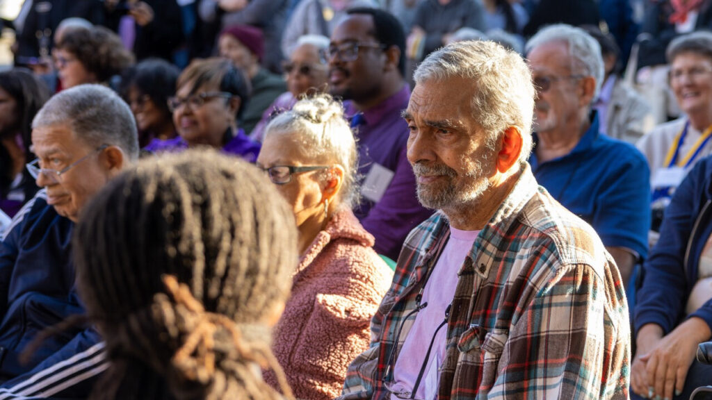 A photo of an older person framed centrally amongst seated people, outdoors in the shade on a sunny day. They are looking an unseen presentation to the left.
