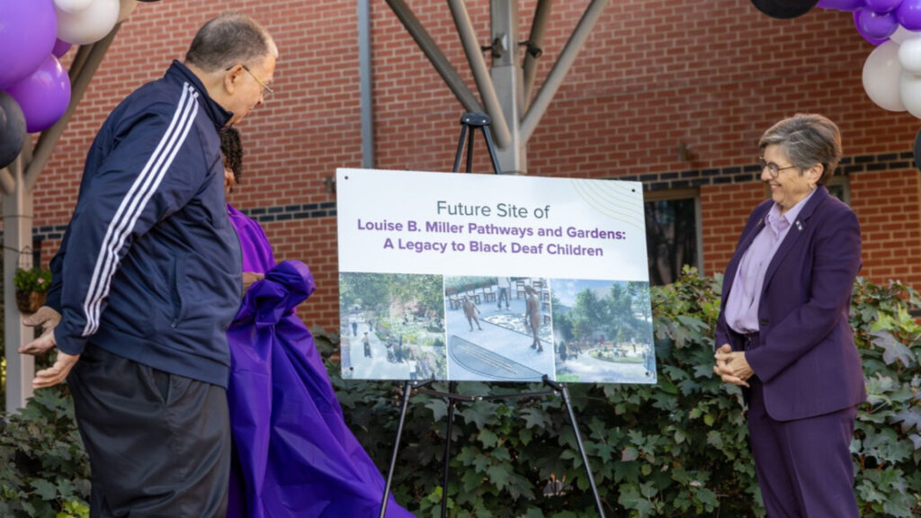 A photo of two people on an outdoor stage looking at an unveiled sign that says, "Future Siate of Louise B. Miller Pathways and Gardens: A Legacy to Black Deaf Children". The sign has three images below the text. The person on the right is smiling.