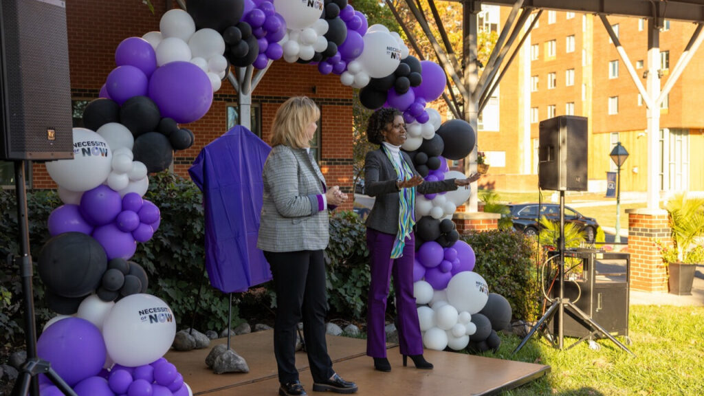 A photo of two people on an outdoor stage presenting to an unseen crowd to the right. Behind them on the stage is an arc of white, purple, and black balloons, as well as a sign with a purple cloth covering it. Sound system speakers stand on both sides of the stage.