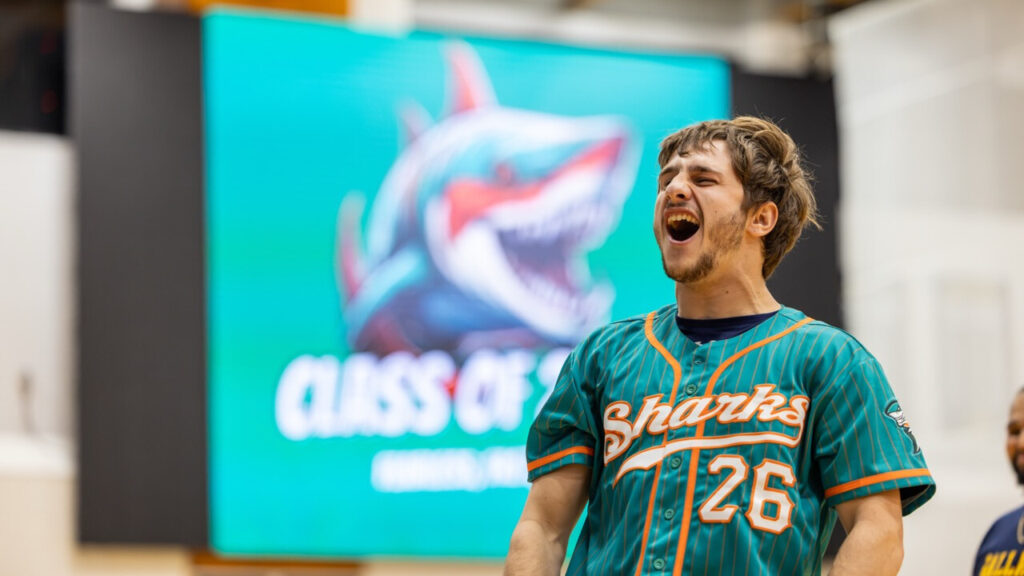 A photo of a college student in a school gym, with an out-of-focus screen in the background that can reads "Class of" with the number obscured. The student is wearing a blue-green baseball jersey that reads, "Sharks ’06". They are shouting intensely to an unseen crowd to the left.