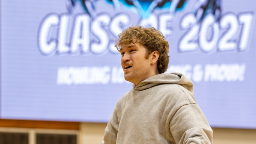 A photo of a college student in a school gym, with an out-of-focus screen in the background that can reads "Class of 2027". The student is emoting vividly to an unseen crowd to the left.