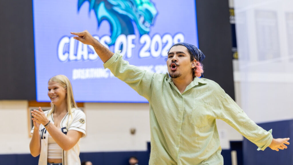 A photo of a college student in a school gym, with an out-of-focus screen in the background that can be assumed to read "Class of 2028". The student is signing enthusiastically to an unseen crowd to the left. Another student is out of focus in the bottom left, farther away.