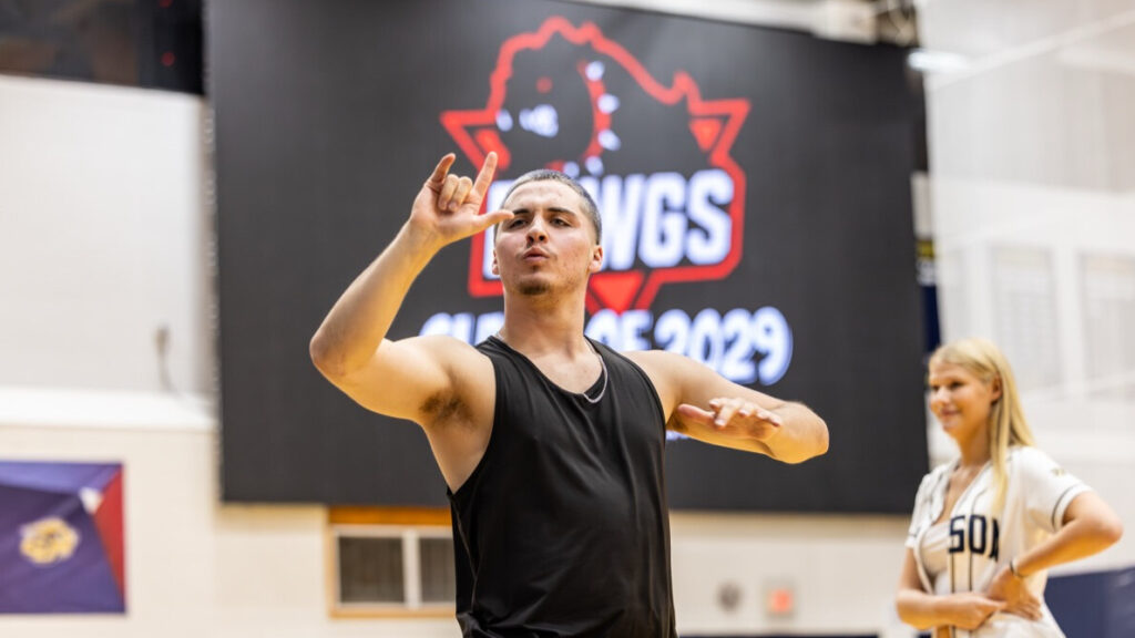 A photo of a college student in a school gym, with an out-of-focus screen in the background that can be assumed to read "Class of 2029". The student is signing to an unseen crowd to the left. Another student is out of focus in the bottom right, farther away.