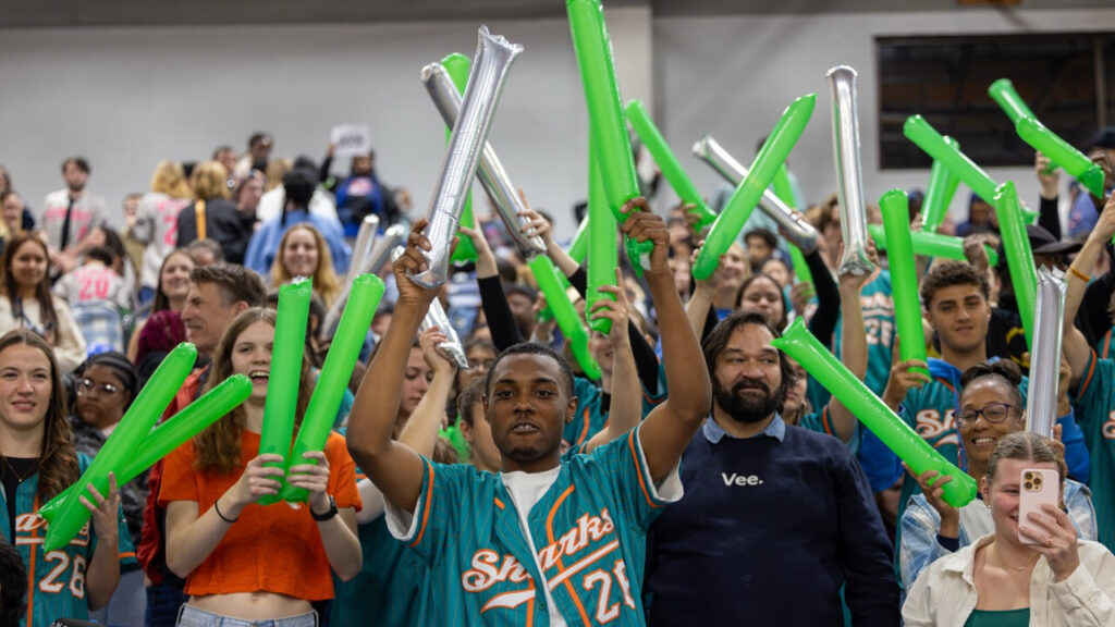 A photo of a lively college pep rally, looking up at a crowd on bleachers. Many of the young people pictured in front are holding up orange and silver thundersticks. Prominent in the bottom center is a smiling person with an unbuttoned blue-green baseball jersey that reads, "Sharks".