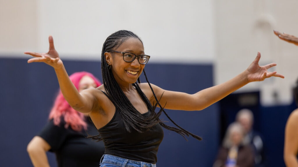 A photo of a dancer in a school gym. They are smiling and holding their arms out wide during a routine. They have glasses and braids over their shoulders. They wear a black tank top. A few people are out of focus in the background.