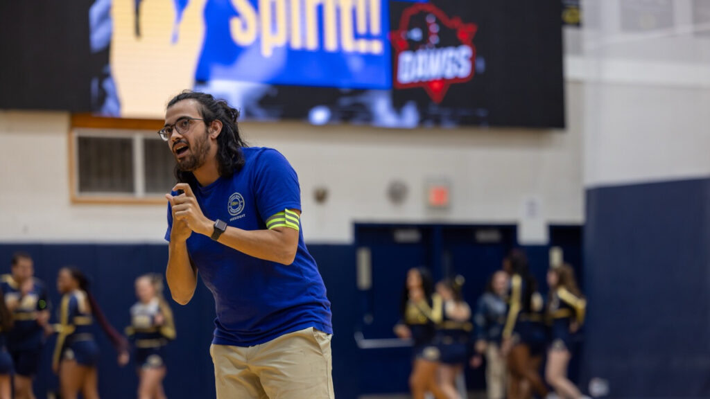 A photo of a person, center left, in a school gym. They are clasping their hands next to their chest as they address an unseen crowd to the left. They wear a blue shirt. In the background, out of focus, are cheerleads and a gym wall.