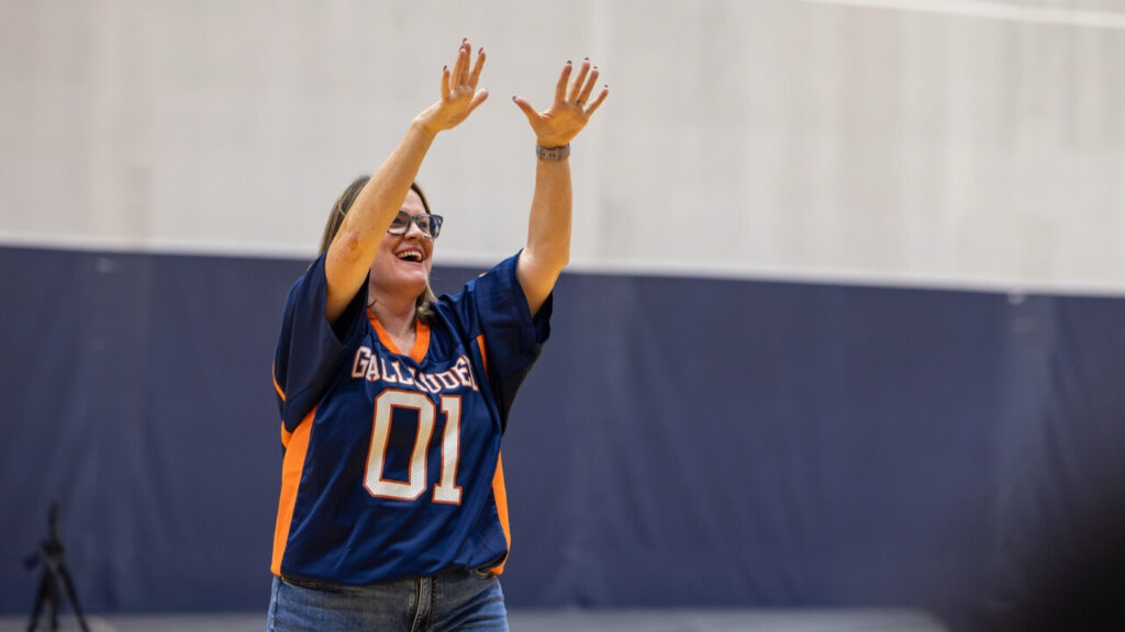 A photo of a person, center left, in a school gym. They are raising their arms in cheer to an unseen crowd to the right. They wear a dark blue and red football jersey that reads, "Gallaudet ’01".