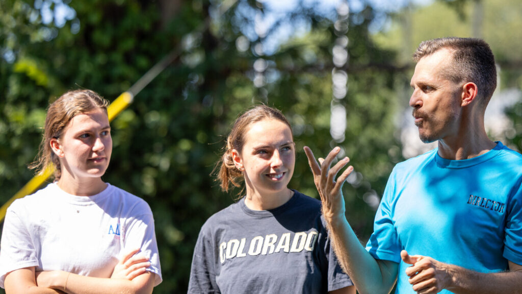 A photo of a middle-aged person, right, signing to two college-aged people on the left, who are watching. The person on the right has a blue t-shirt. The person in the middle has a t-shirt that says "Colorado".