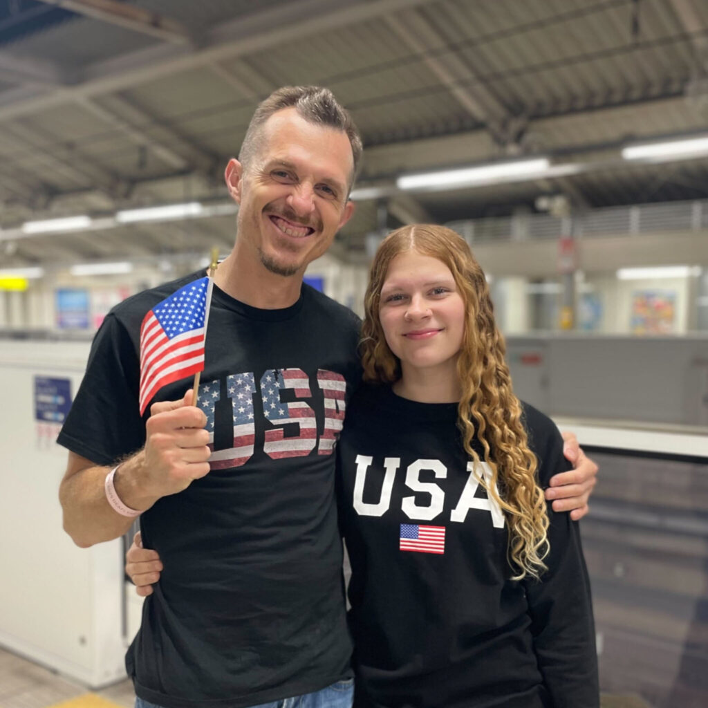 A photo of a father and daughter smiling for the camera together. They wear tops that read "USA" with various USA flag designs. The father on the left is waving a small USA flag. The background is a blurred subway station.