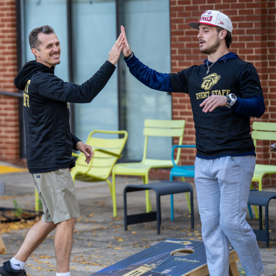 A photo of two people high fiving. The person in the middle of the image is smiling and looking offscreen to the right. The person on the right has a dark blue shirt with the Gallaudet Athletics logo and text: "Event Staff". A cornhole board is below them.